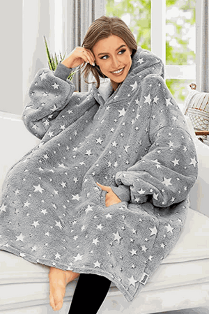 Woman wearing oversized gray Lazy Fleece Blanket Sweater with white stars, sitting indoors and smiling
