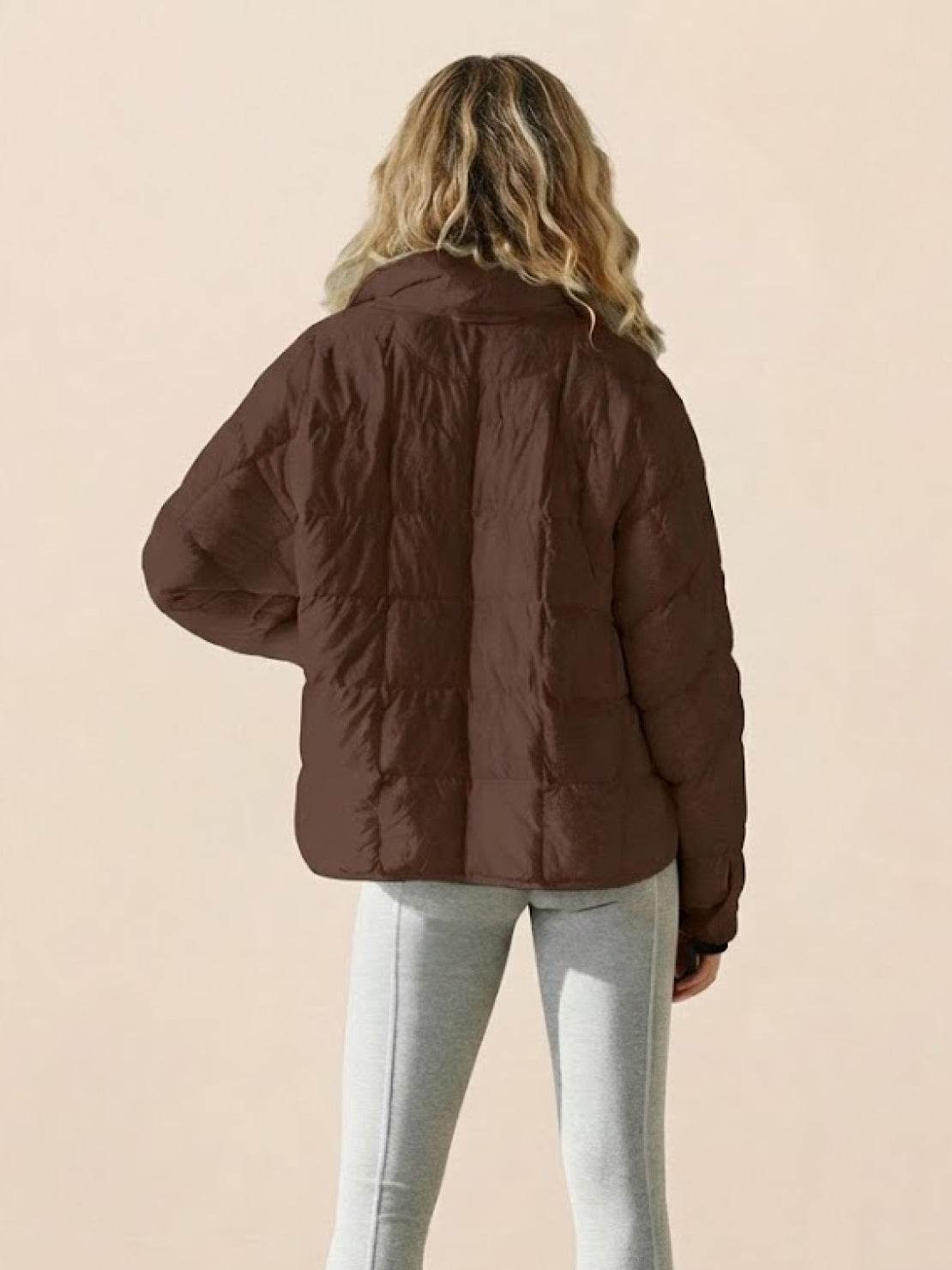 Woman wearing a brown lightweight puffer travel jacket from the back with gray pants against a beige background