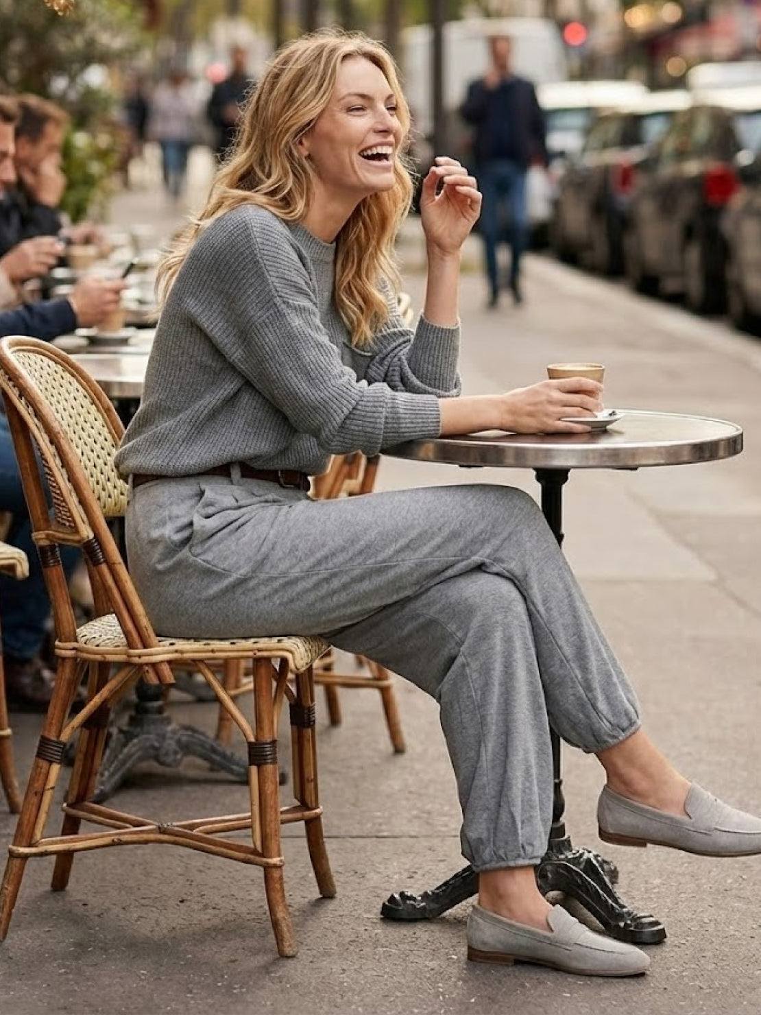 Woman wearing European minimalism gray relaxed sweater top and trouser set sitting at outdoor café table with coffee cup