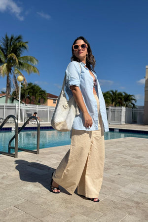 Woman wearing oversized linen shirt with beige wide-leg pants by poolside under clear blue sky