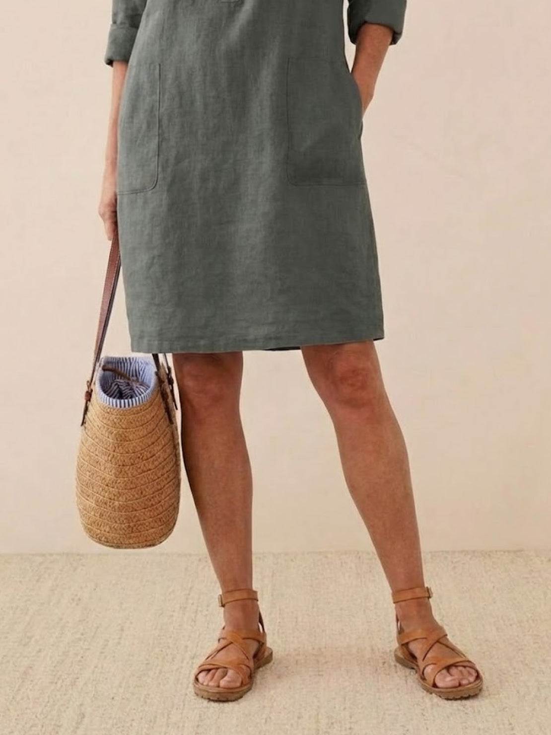 Woman wearing knee-length gray cotton dress with 3/4 rolled sleeves, holding straw handbag and brown sandals.
