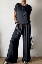 Woman wearing Silken Escape satin black two-piece set with wide-leg pants and short-sleeve top, accessorized with necklace and handbag.