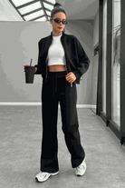 Woman wearing The Voyager Zip Set black zip-front sweatshirt and wide-leg joggers with white sneakers and sunglasses indoors.