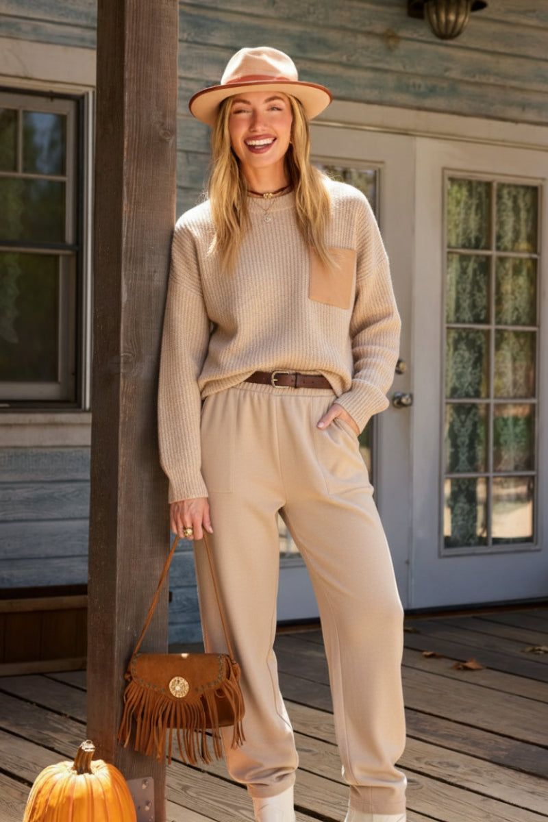 Woman wearing European minimalism relaxed sweater top and trouser set in beige standing on wooden porch with hat and handbag.