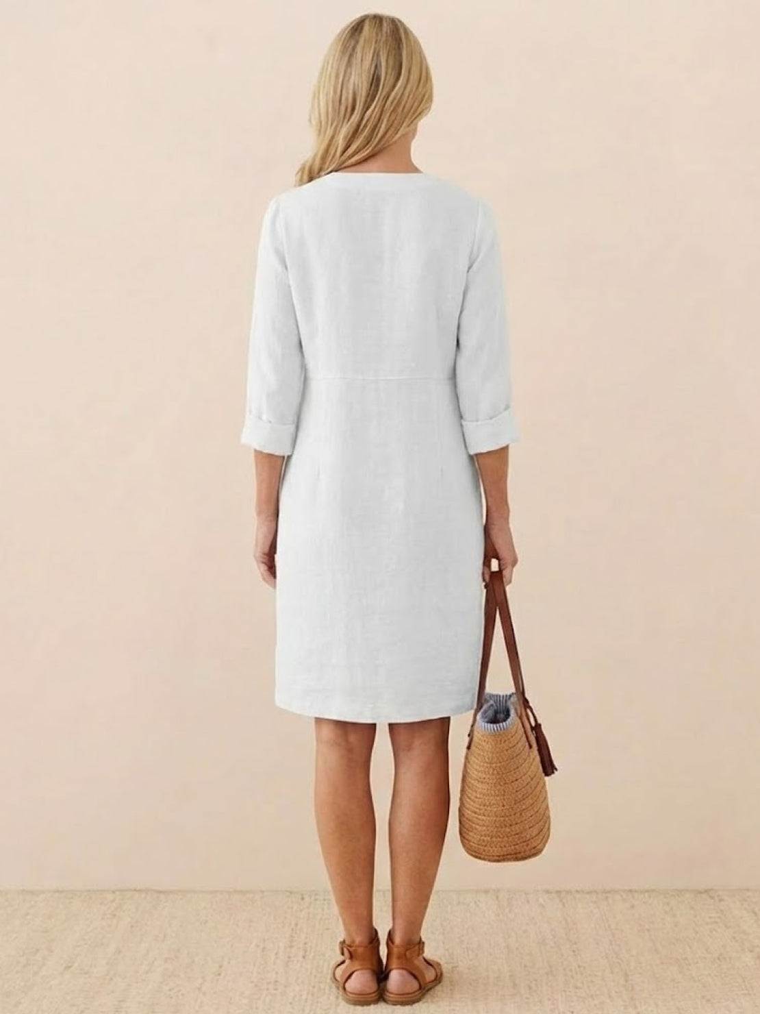 Back view of a woman wearing a simple white cotton dress with 3/4 rolled sleeves, holding a woven handbag, standing against a neutral background.